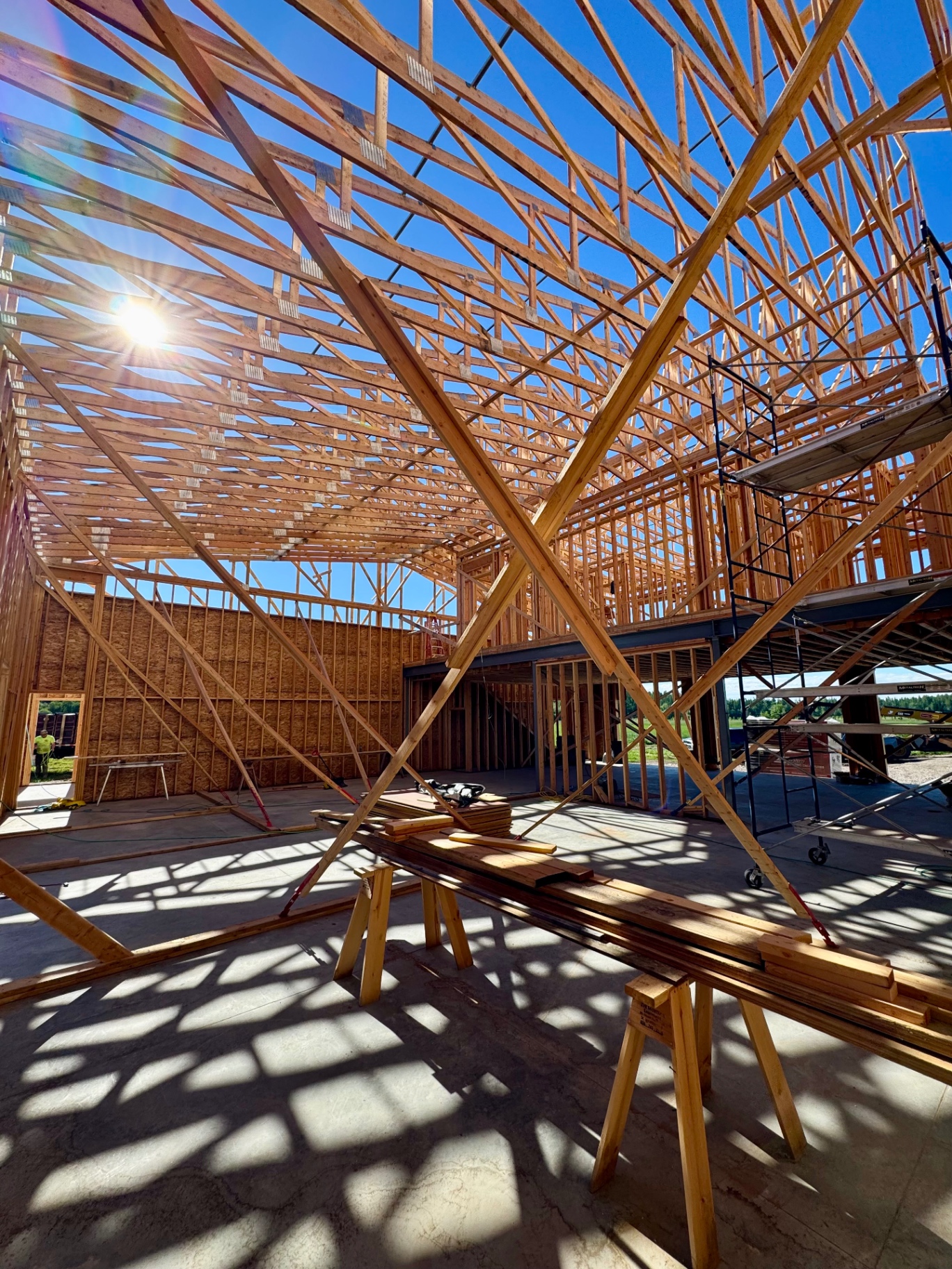 Exposed timber framing and roof trusses inside a custom home under construction in the Flathead Valley, scaffolding and sawhorses silhouetted against a bright blue sky.