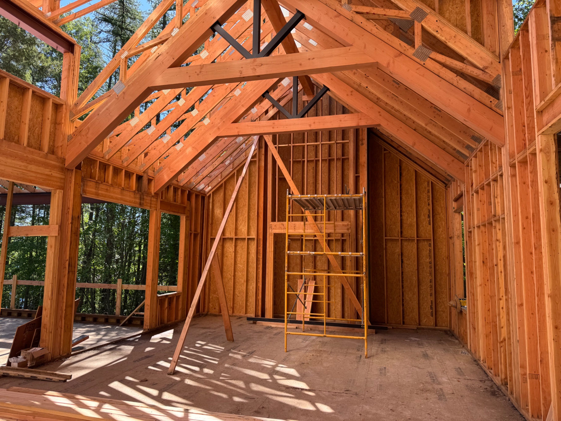 Interior timber framing of a custom home under construction, with exposed Douglas fir trusses, steel tie rods, and a rolling scaffold set against forested Flathead Valley views.